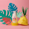 A bottle of Dabble & Dollop Dole Whip® Pineapple Foaming Body Wash sits on a pink background, surrounded by colorful paper leaves and a paper pineapple.