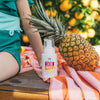 A child in a green swimsuit sits on a pink and orange striped towel beside a fresh pineapple, Dabble & Dollop Dole Whip® Pineapple Foaming Body Wash, and a bottle of pineapple-scented hand soap outdoors with trees and oranges behind them.