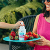 Outdoors among greenery, one person holds a plate of fresh strawberries while another dispenses Dabble & Dollop's Dole Whip® Strawberry Foaming Body Wash, evoking the refreshing scent of summer and natural ingredients.