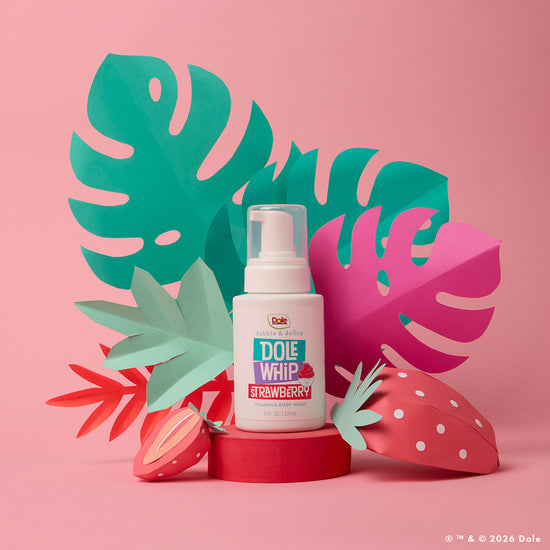 A bottle of Dabble & Dollop Dole Whip® Strawberry Foaming Body Wash sits among vibrant paper cutouts of tropical leaves and strawberries on a pink backdrop, adding a hypoallergenic touch of the tropics to your bath routine.