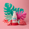 A bottle of Dabble & Dollop Dole Whip® Strawberry Foaming Body Wash sits among vibrant paper cutouts of tropical leaves and strawberries on a pink backdrop, adding a hypoallergenic touch of the tropics to your bath routine.