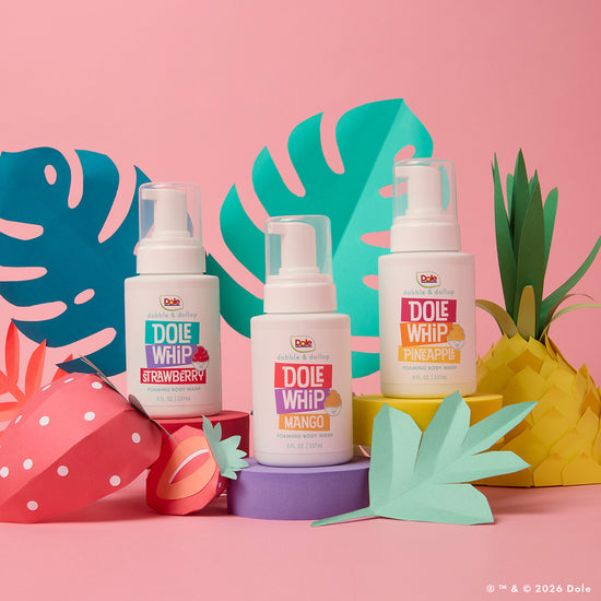 Three bottles of Dabble & Dollop’s Dole Whip® Pineapple Foaming Body Wash—plus strawberry and mango variants—are displayed on a pink background with fruit and leaf cutouts, showcasing their natural-inspired fragrances.
