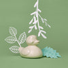 A cream-colored Ducky Bath Toy & Teether by Dabble & Dollop, made in the USA and latex-free, sits on a green surface with white and light blue paper leaves, against a matching green background.