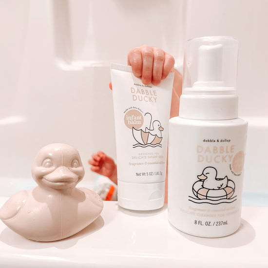 A childs hand holds a tube of infant balm in a bathtub, with a latex-free foaming cleanser bottle nearby. A cheerful Dabble & Dollop Ducky Bath Toy & Teether sits on the tubs edge. Made in the USA.