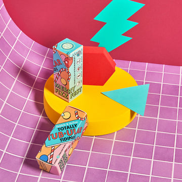 Two brightly decorated rectangular boxes of Dabble & Dollop’s Totally Tub-ular Merry Fizzfest Mini Drops Stocking Stuffer perch on colorful shapes against a vivid pink grid, making a playful holiday gift or perfect stocking stuffer!.