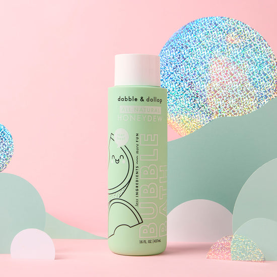 A green bottle of Dabble & Dollop Honeydew Bubble Bath stands upright against a pastel pink background with holographic and pastel paper cutout hills and clouds, emphasizing its gentle, kid-friendly formula.