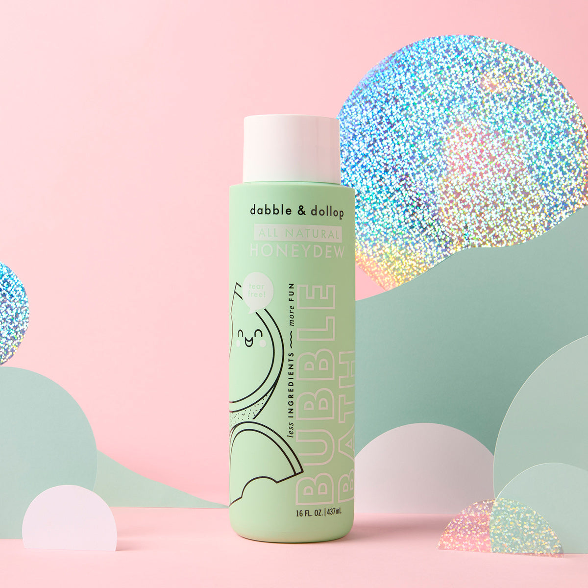 A green bottle of Dabble & Dollop Honeydew Bubble Bath stands upright against a pastel pink background with holographic and pastel paper cutout hills and clouds, emphasizing its gentle, kid-friendly formula.