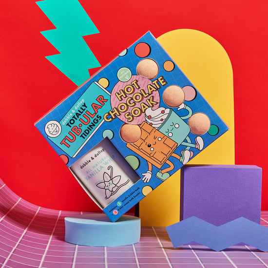 A vibrant display shows Dabble & Dollop’s Totally Tub-ular Tidings Bubble Bath Set—featuring playful cartoon graphics, hot chocolate-scented bath bombs, and a vanilla candle—set against a bold geometric red, yellow, and blue background.