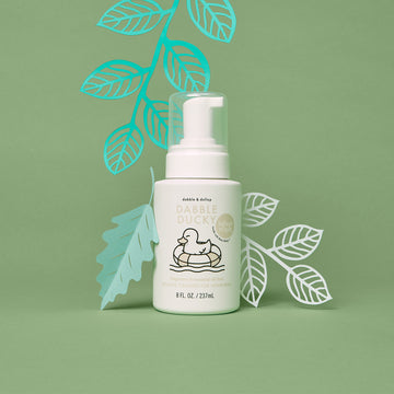 A bottle of Dabble & Dollop Fragrance Free Foaming Infant Wash, perfect for sensitive skin, is displayed against a green backdrop with paper-cut leaves. This natural baby wash features duck art and contains 8 fl. oz.