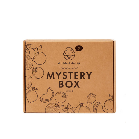 A brown cardboard box labeled Dabble & Dollop Mystery Bath Bundle features fruit illustrations around the edges and a smiling face with a question mark—packed with bubbly creativity and delightful bath surprises inside.
