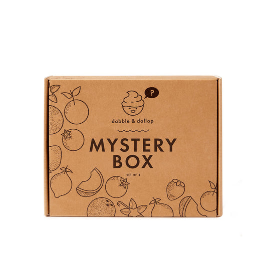 A brown cardboard box labeled Dabble & Dollop Mystery Bath Bundle features fruit illustrations around the edges and a smiling face with a question mark—packed with bubbly creativity and delightful bath surprises inside.