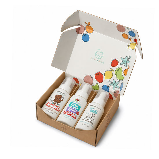 An open cardboard box reveals three Dabble & Dollop Bath Gift Sets—ideal for kids’ bath time or as a thoughtful gift—featuring colorful fruit illustrations on the inside of each lid. Each bottle is labeled with one of the following scents: Candy Cane Chocolate, Dole Whip Strawberry, and Dole Whip Pineapple. The gentle formulas are specially designed for sensitive skin.