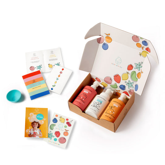 The Dabble & Dollop Original Bubble Bath Bundle features an open box with three kids’ bath products, colorful cards, a blue cup, and playful fruit-themed packaging—all made with clean ingredients.