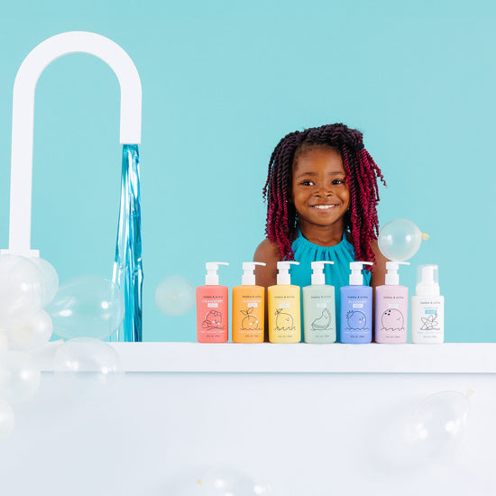 A smiling child with braided hair stands behind a counter with six colorful bottles from the Dabble & Dollop Rainbow of Bubbles Bath Bundle, as bubbles float around and water pours near a bright turquoise background.