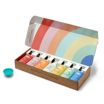 The Dabble & Dollop Rainbow of Bubbles Bath Bundle features six colorful bubble bottles in matching pastels, arranged in a box with an open lid. A blue bubble wand sits next to the set, perfect for bath time fun.