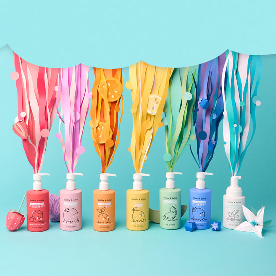 Dabble & Dollops Rainbow of Bubbles Bath Bundle features a row of vibrant body wash and shampoo bottles with fruit designs, topped by matching paper streamers and fruits in rainbow colors, set against a blue backdrop.