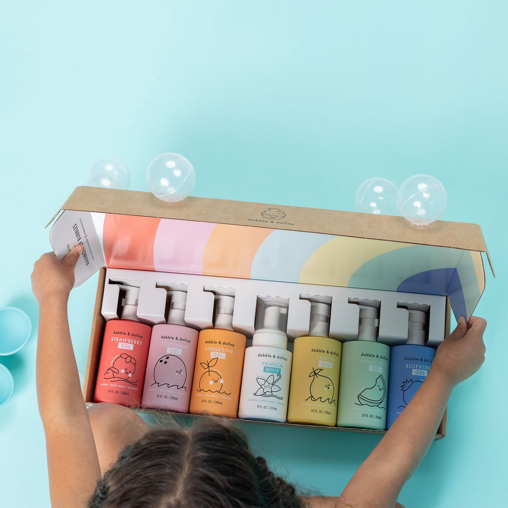 A child holds an open box of Dabble & Dollop’s Rainbow of Bubbles Bath Bundle—six colorful bottles (shampoos, body wash & bubble bath) with playful line art, arranged in a row against a bubble-filled light blue background.