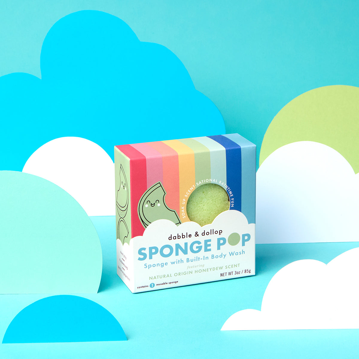 A box of Dabble & Dollop Sponge Pop™ Sponge with Built-In Body Wash features rainbow stripes, a cartoon character, and text, against a colorful background with blue and white paper clouds. This sensory bath toy has a honeydew scent.