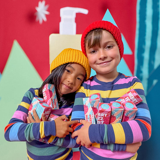 Two children in colorful striped shirts and knit hats hug each other, smiling and holding several boxes of the Dabble & Dollop Merry Fizzfest Mini Drops Stocking Stuffer—a cheerful holiday gift filled with festive bath bombs. The bright background is adorned with holiday-themed decorations.