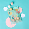 A turquoise and pink flat lay background features a bag of Dabble & Dollop Tropical Mini Bath Bombs, surrounded by round tropical bath bombs and white and pink paper circles.