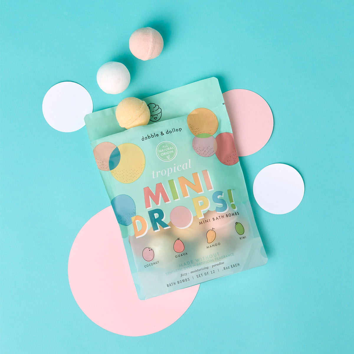 A turquoise and pink flat lay background features a bag of Dabble & Dollop Tropical Mini Bath Bombs, surrounded by round tropical bath bombs and white and pink paper circles.