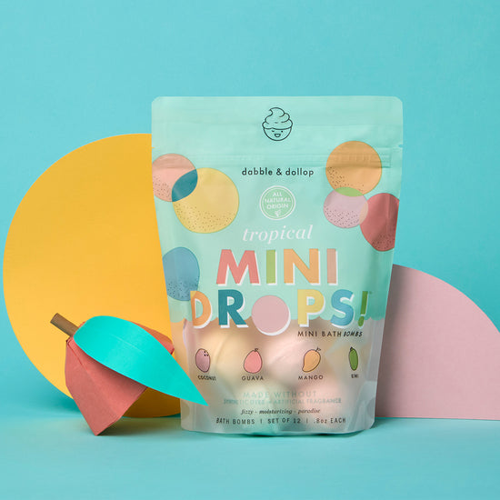 A colorful pack of Dabble & Dollop Tropical Mini Bath Bombs for kids is set against a blue background with pastel yellow and pink paper shapes and a decorative paper fruit, featuring illustrated fruit flavors on the packaging.