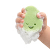 A hand squeezes Dabble & Dollop's Sponge Pop™ Sponge with Built-In Body Wash, a green biodegradable sponge with a winking smile, covered in foamy bubbles against a white background.
