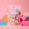 A pastel pouch of Dabble & Dollop Sweet Treat Mini Bath Bombs stands upright on a pink surface, surrounded by colorful paper cutouts and two pink gift boxes. The playful packaging suggests dessert-inspired bath bombs for sensitive skin.