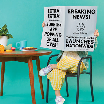 Child sitting at toy kitchen table setup and holding up and reading a newspaper